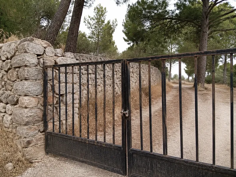 Portalón metálico y muro de piedra en un camino, cerradura de cancela en primer plano, sin personas
